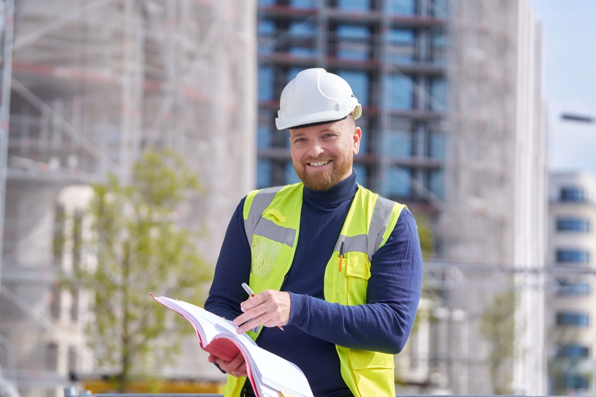 Alexander Michel mit Schutzhelm und Warnweste auf einer Baustelle, in der Hand eine Projektmappe